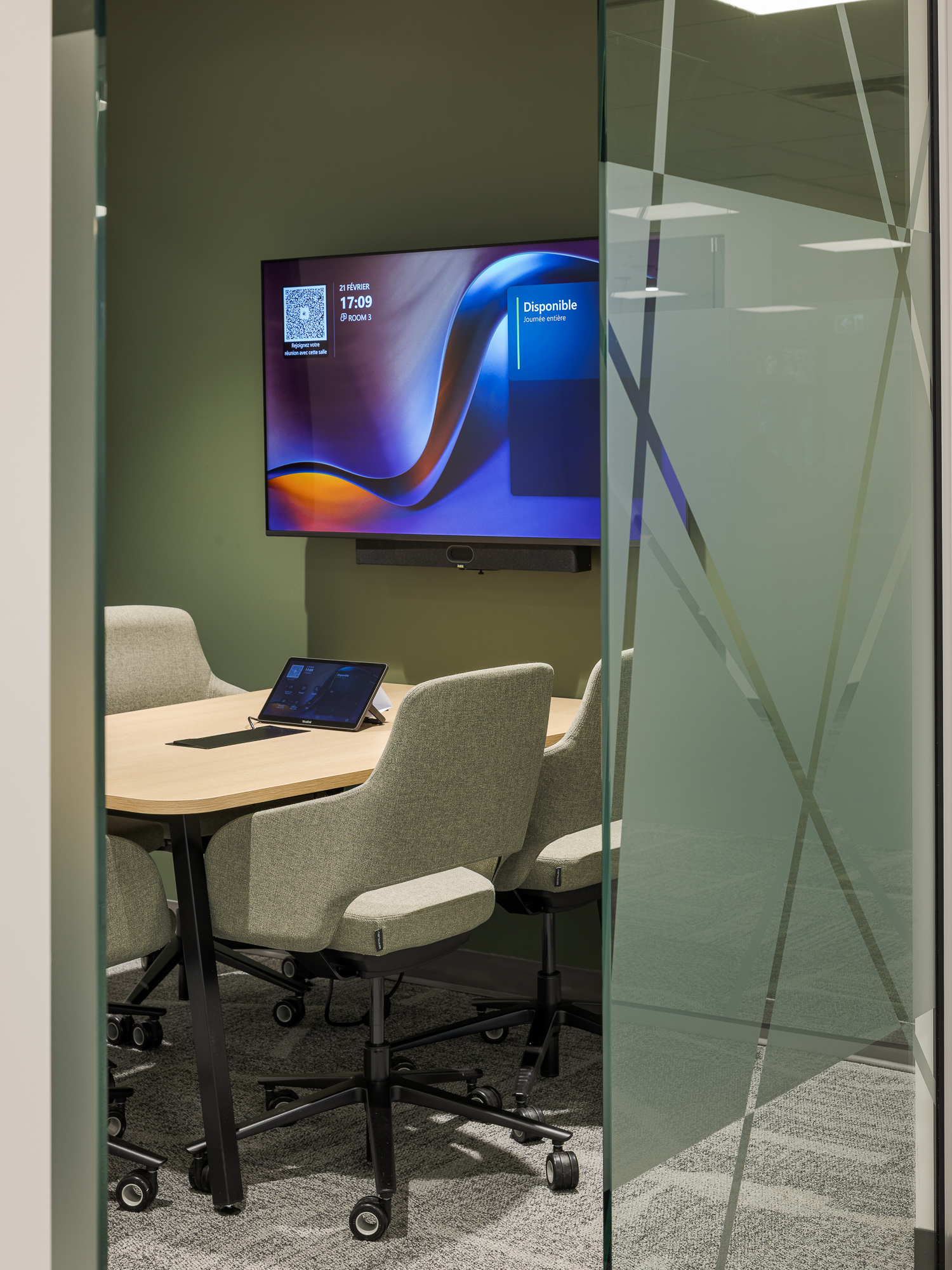 A modern office meeting room designed by Aquest Design, featuring sage green walls, glass window, collaborative tech integration, and stylish grey upholstered seating.